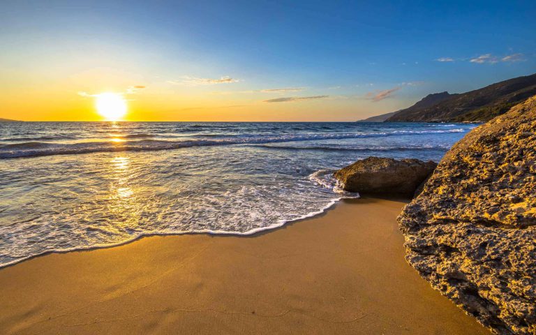 Sonnenuntergang über einem Strand mit sanften Wellen und felsiger Küste.