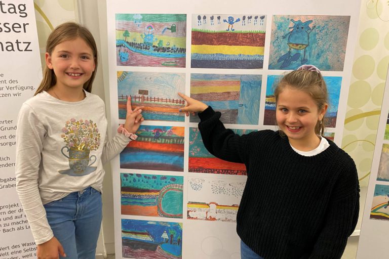 Zwei Mädchen zeigen stolz ihre kunstvollen Bilder zur Wanderausstellung über Wasser.