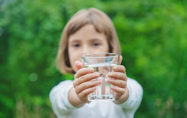 Kind hält ein Glas Wasser vor grünem Hintergrund, symbolisiert gesunde Trinkgewohnheiten.